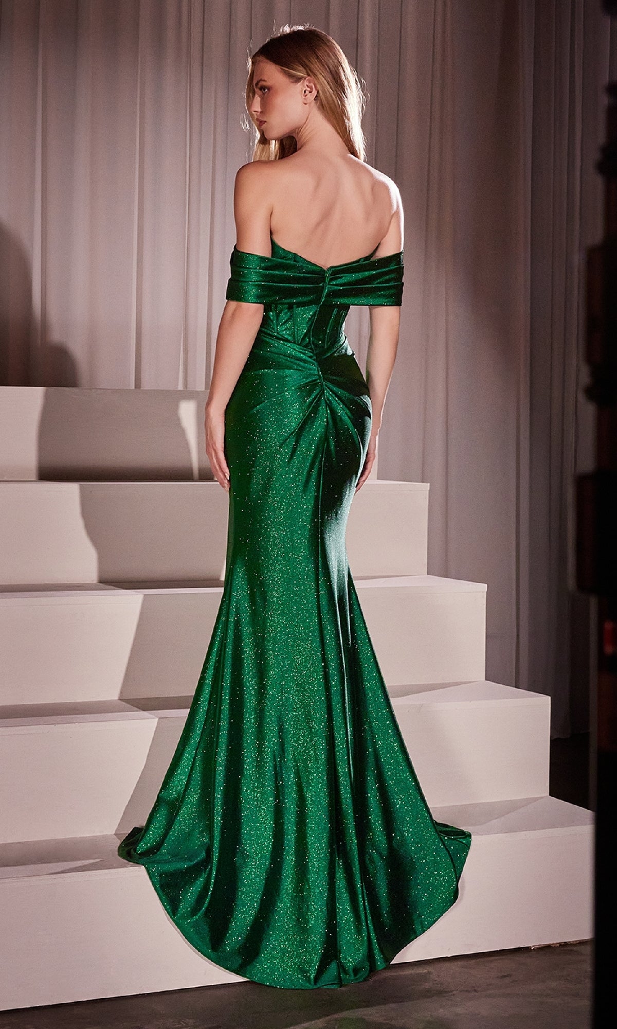 A woman faces away on white steps, wearing the Ladivine KV1115—a floor-length, emerald green glitter satin prom dress with off-the-shoulder sleeves, fitted silhouette, slight train, and subtle shimmer, perfect for special occasions.